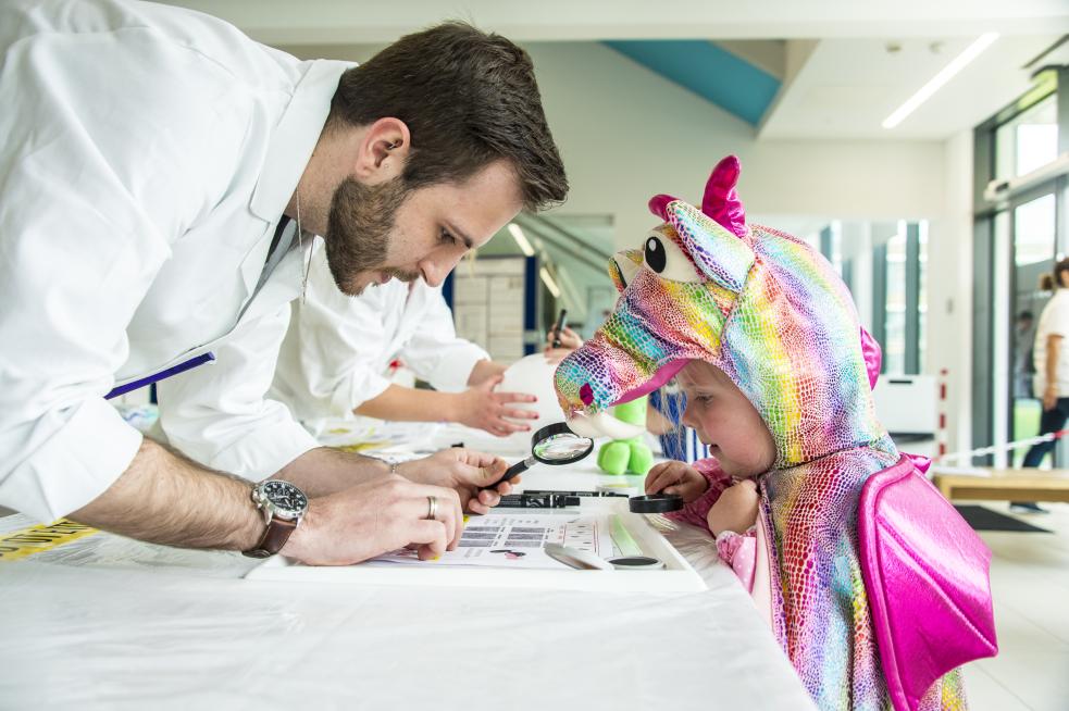 man in lab coat looking at something through a magnifying glass with younger child dressed in a.multicoloured horse outfit