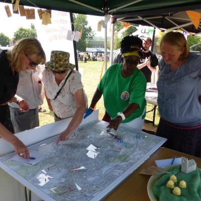  A group of people standing around a table putting pins on a large map, at an ourdoors event in the sunshine