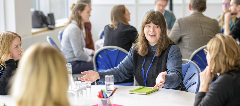 4 people round a table talking and gesturing with similar group discussions faded in background