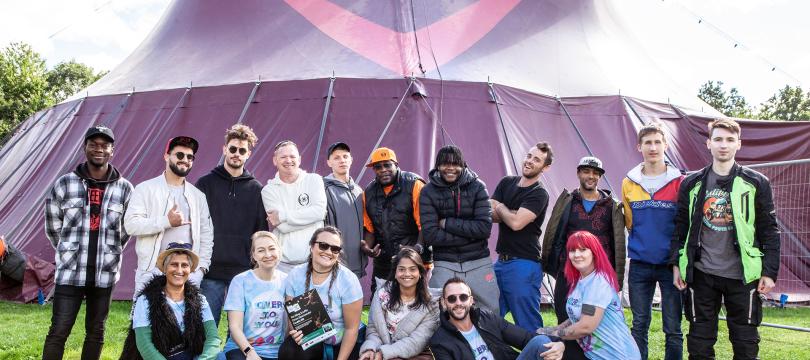 group of young people standing and sitting and smiling in front of a bit purple tent outside