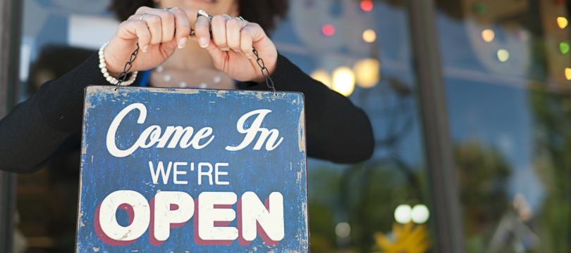 A woman smiling and holding up a shop sign that says 'come in we're open'