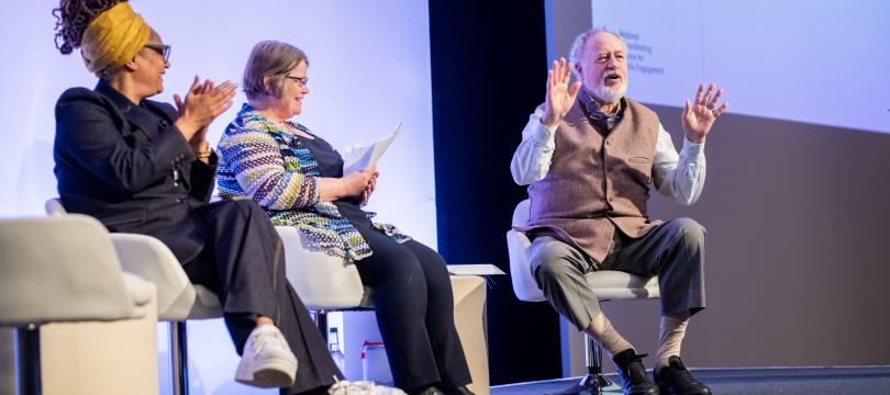 Budd Hall sitting, talking animatedly on stage at Engage with Sophie Duncan and Ima Jackson 