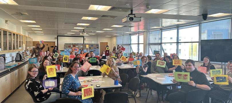 A classroom of people sat at tables holding up printed artwork