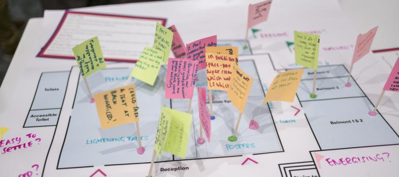 A floor plan of a conference venue overlaid with small handwritten notes on coloured paper flags stuck into the plan with cocktail sticks. The notes capture participant feedback and observations at different points in the space, including areas labelled Plenary, Lightning Talks, Posters, Reception, Arriving and Entrance. The overall effect is of a participatory mapping or experience evaluation exercise conducted during or after the event.