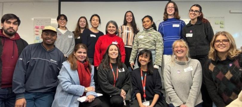 A group of 15 in-person attendees smiling together in a workshop setting – some kneeling in the front row and others standing in the back in front of a white screen.