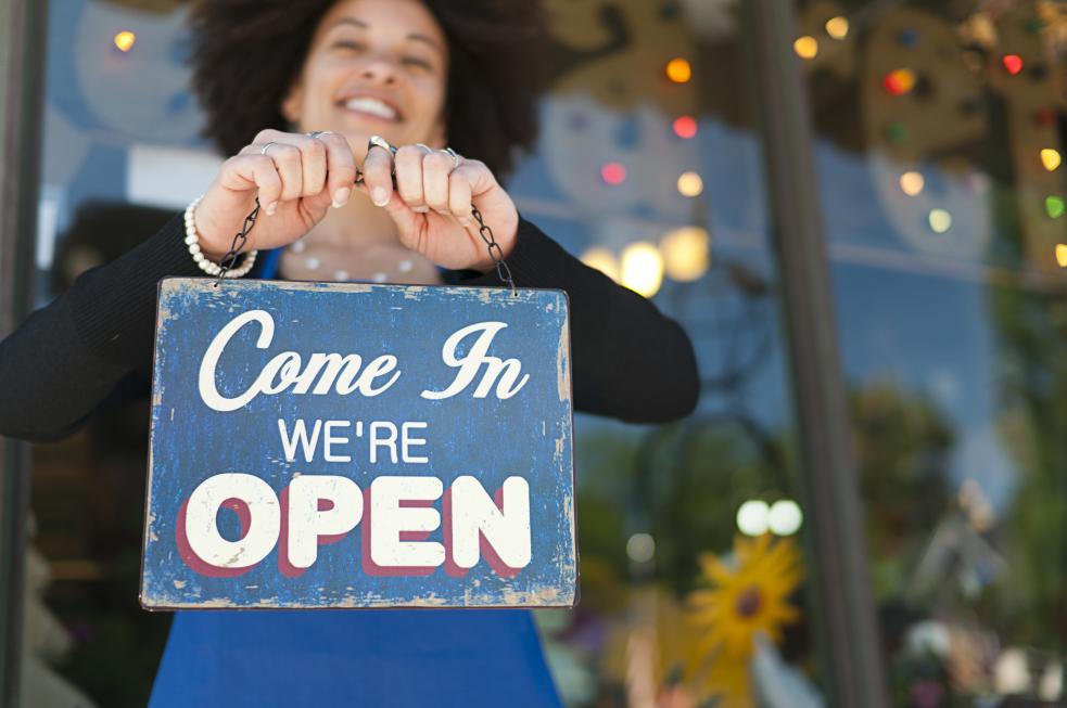 A woman smiling and holding up a shop sign that says 'come in we're open'