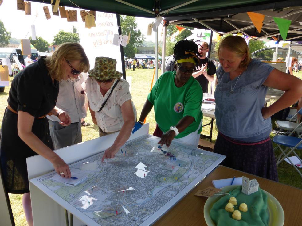 A group of people standing around a table putting pins on a large map, at an ourdoors event in the sunshine