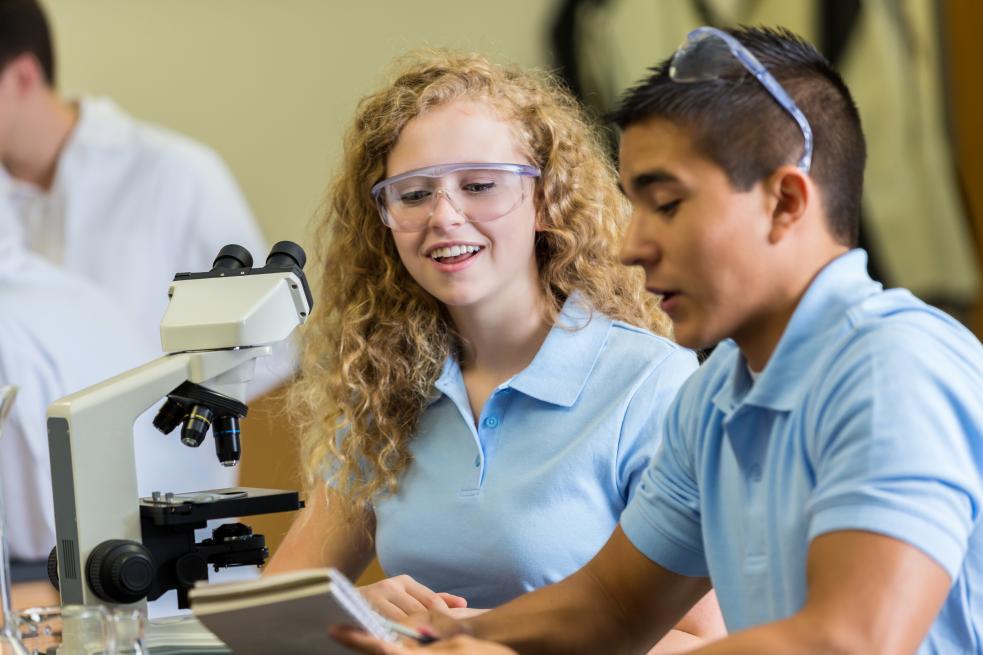Two young people sit on a table with a microscope. They are discussing something together and smiling