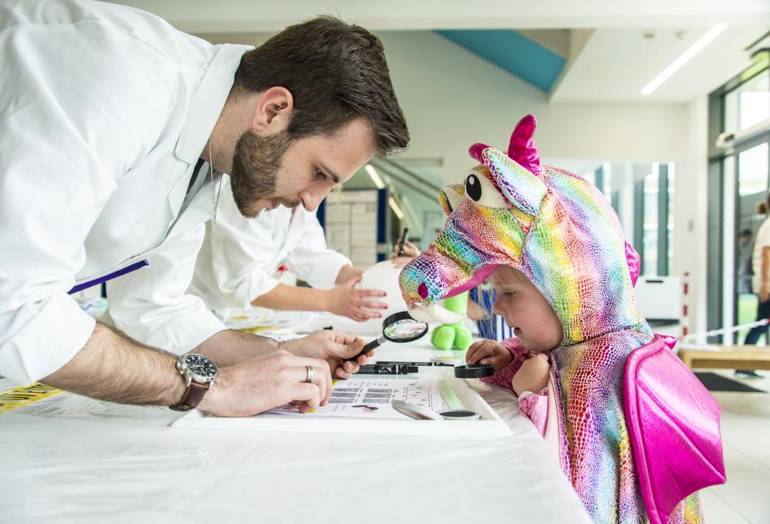 man in lab coat looking at something through a magnifying glass with younger child dressed in a.multicoloured horse outfit