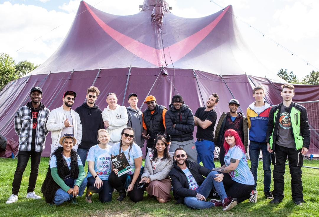 group of young people standing and sitting and smiling in front of a bit purple tent outside