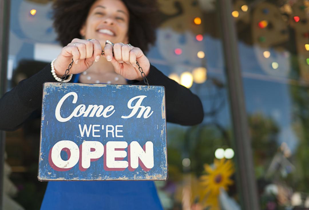 A woman smiling and holding up a shop sign that says 'come in we're open'