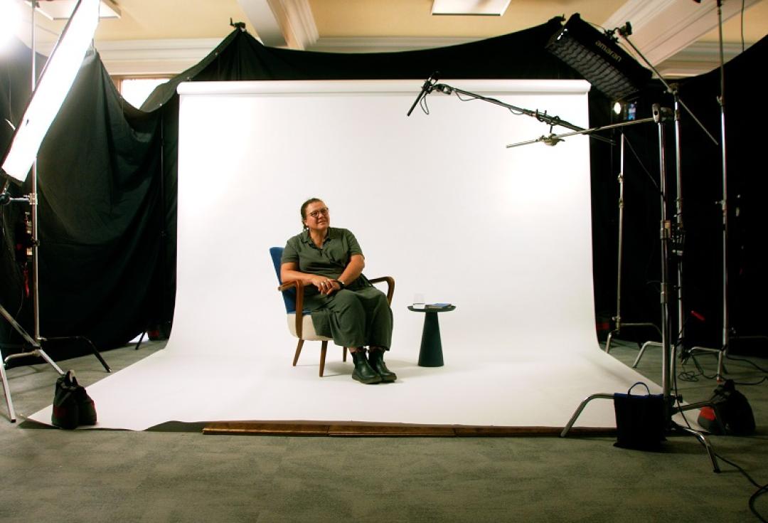 Researcher in a recording studio sat in a chair in front a screen with microphones ready to be filmed