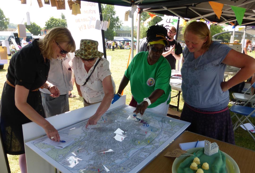  A group of people standing around a table putting pins on a large map, at an ourdoors event in the sunshine