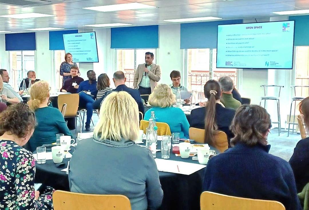 Ayad presenting to a group of about 25 people sitting in chairs in a meeting room. There are 2 large screens in the background with a presentation on