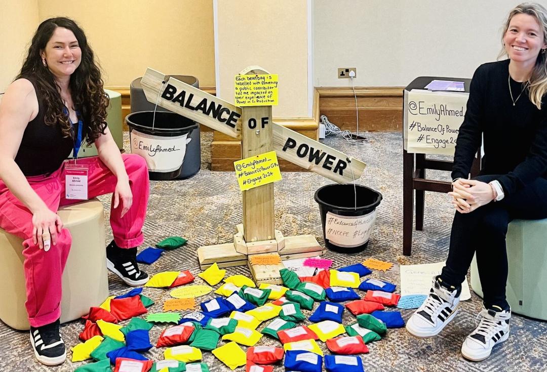 Emily and her colleague sitting on stools either side of Emily's 'balance of power' wooden scales, surrounded by colourful beanbags with labels attached.