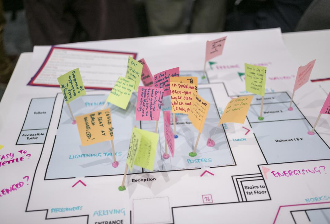 A floor plan of a conference venue overlaid with small handwritten notes on coloured paper flags stuck into the plan with cocktail sticks. The notes capture participant feedback and observations at different points in the space, including areas labelled Plenary, Lightning Talks, Posters, Reception, Arriving and Entrance. The overall effect is of a participatory mapping or experience evaluation exercise conducted during or after the event.