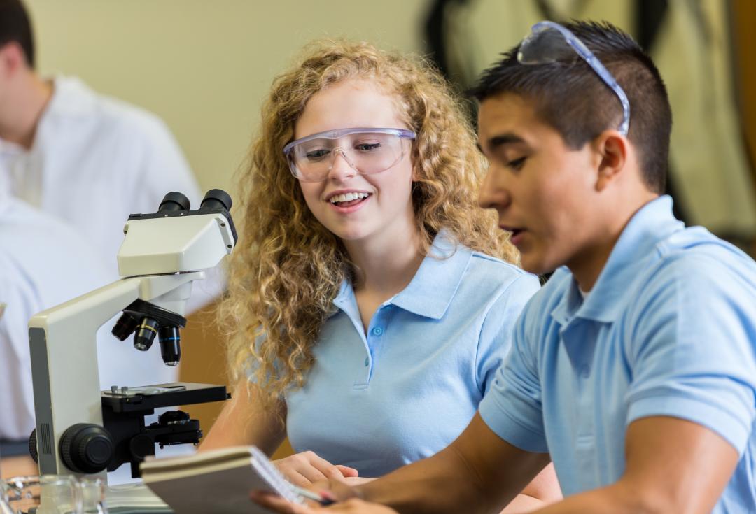 Two young people sit on a table with a microscope. They are discussing something together and smiling