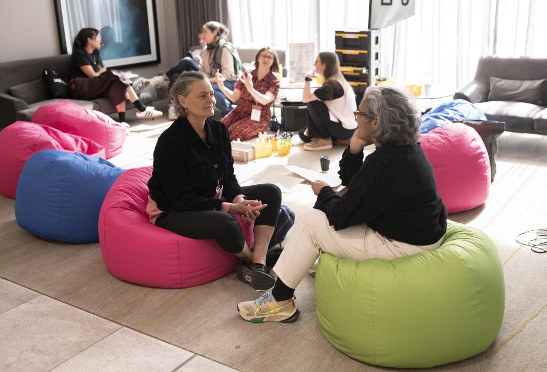 Two delegates seated in pink and green beanbags in conversation, holding papers, with other small groups talking on sofas and beanbags in a bright room behind them.