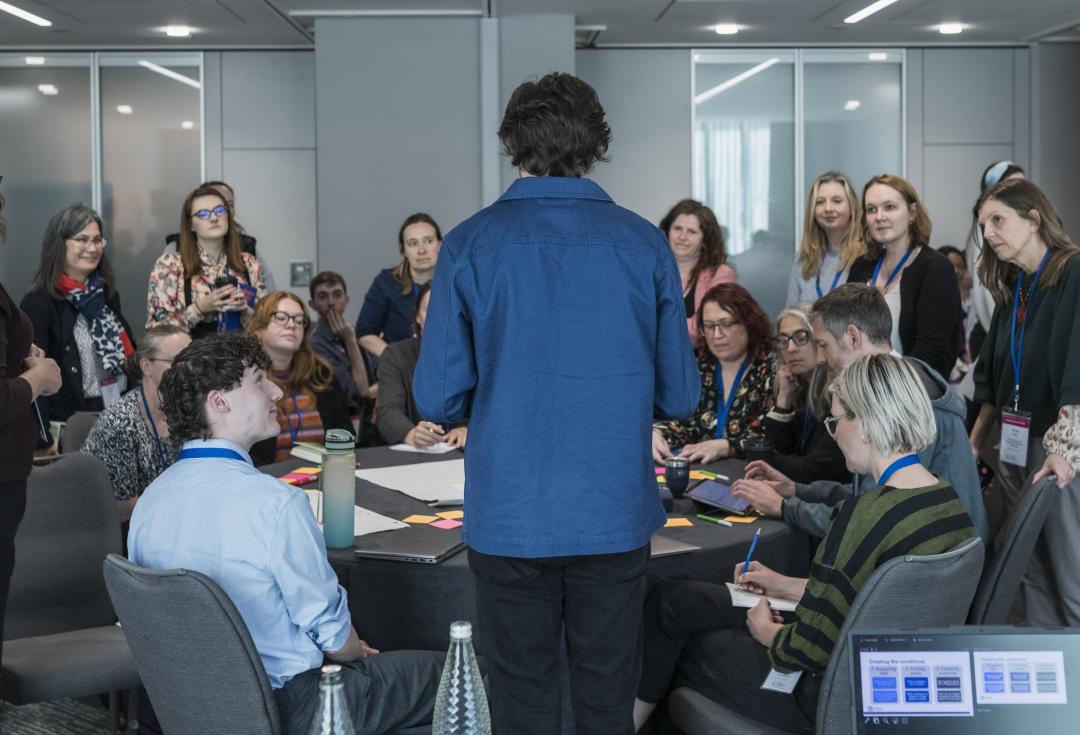 A group of people are sitting at a table and listening a colleague explain something