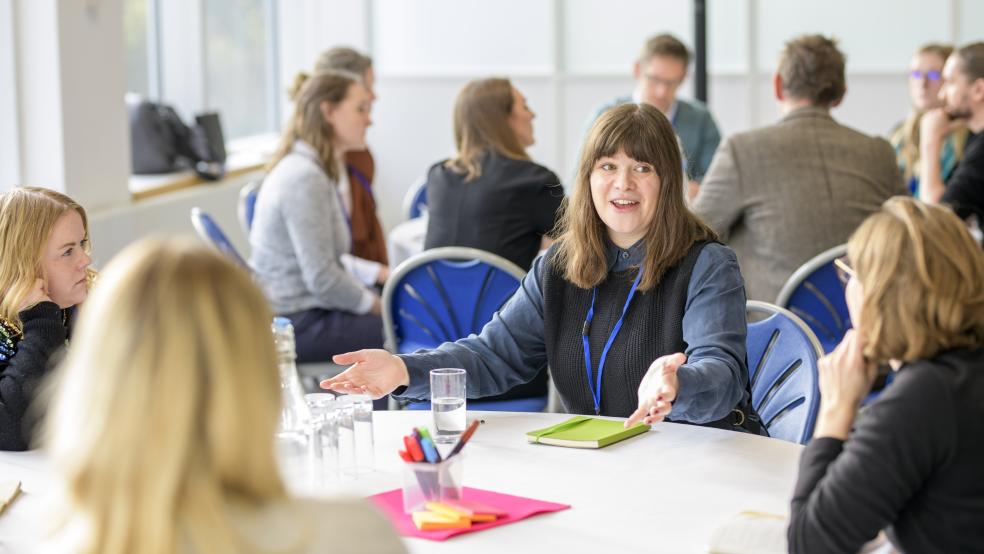 4 people round a table talking and gesturing with similar group discussions faded in background