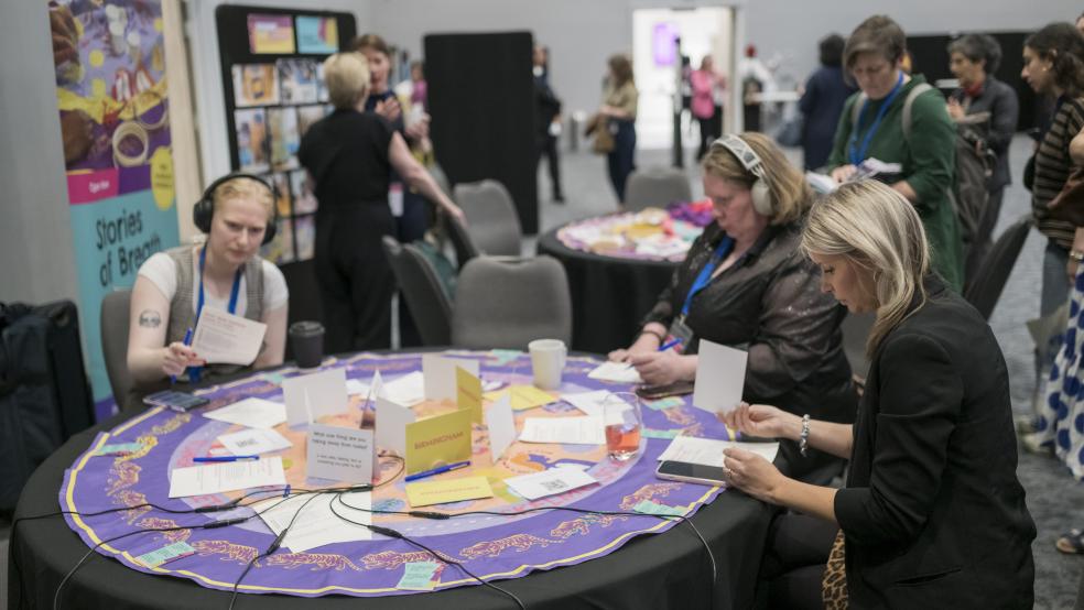 Three women sit at a round table. Two are wearing headphones l