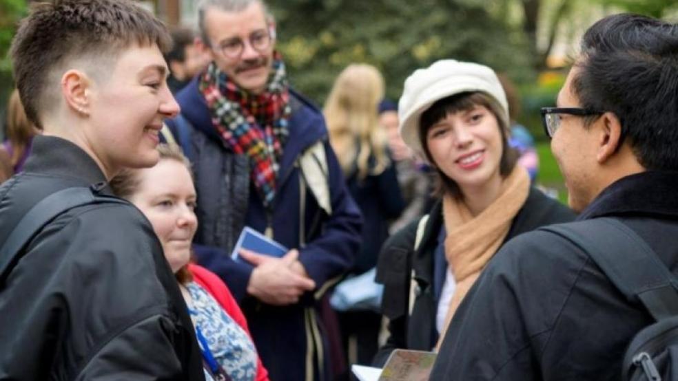 A group of people stand talking outdoors in a park or garden, with trees and a brick building behind them. Five people are gathered in the foreground in conversation, some smiling, with more people visible in the background. Several are wearing coats and scarves, suggesting cool weather, and a couple are holding leaflets or booklets.