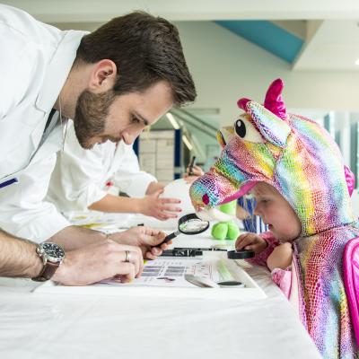 man in lab coat looking at something through a magnifying glass with younger child dressed in a.multicoloured horse outfit