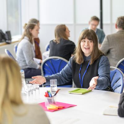 4 people round a table talking and gesturing with similar group discussions faded in background