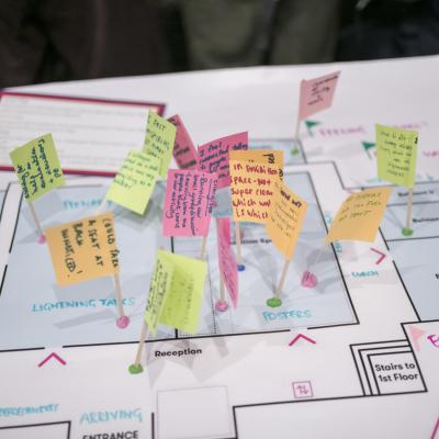 A floor plan of a conference venue overlaid with small handwritten notes on coloured paper flags stuck into the plan with cocktail sticks. The notes capture participant feedback and observations at different points in the space, including areas labelled Plenary, Lightning Talks, Posters, Reception, Arriving and Entrance. The overall effect is of a participatory mapping or experience evaluation exercise conducted during or after the event.