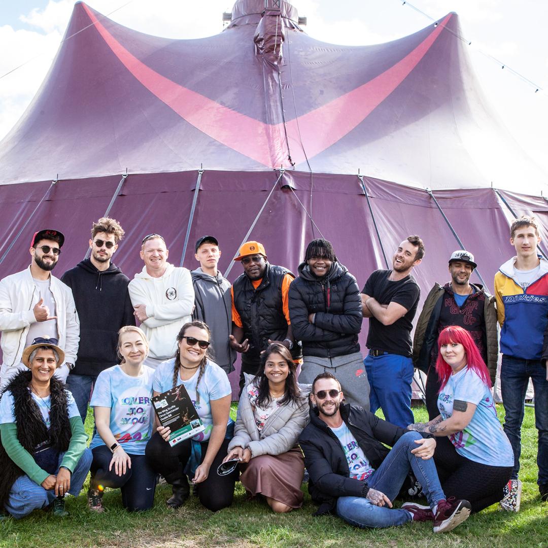 group of young people standing and sitting and smiling in front of a bit purple tent outside