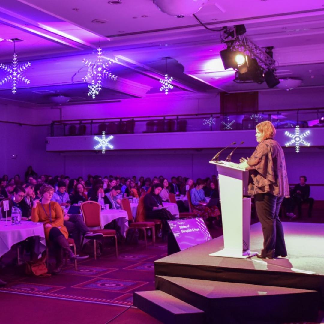 A woman stands on stage presenting to an auditorium of delegates